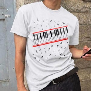 May include: White t-shirt featuring a piano keyboard design with black and white keys and red accents. Musical notes are scattered around the keyboard graphic. The shirt has short sleeves and a crew neck.