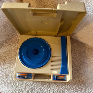 May include: Vintage beige and blue portable record player with a hinged lid. The turntable has a blue platter and a blue arm. The control panel has orange and blue accents. The record player is open and ready to play records.