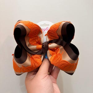 May include: A large hair bow featuring a layered design of tan satin ribbon and dark brown grosgrain ribbon. The bow is decorated with orange pumpkin-shaped fabric appliques along the edges and is secured with a central dark brown knot.