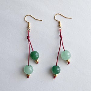 May include: A pair of drop earrings featuring gold-toned fishhook wires, thin maroon cord strands, and two round green stone beads on each earring. Each bead is accented with a small gold-toned metal spacer bead at the bottom of the cord.