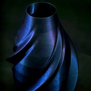 May include: A dark blue, iridescent vase with a swirling, sculptural design. The vase has a wide, flared base that tapers to a narrow opening at the top. The surface reflects shades of blue and purple.