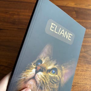 May include: A personalized notebook featuring a close-up image of a cat's face with blue and green eyes. The name "ELIANE" is printed in gold on the cover. The text "Images may show a sample name" is also visible.