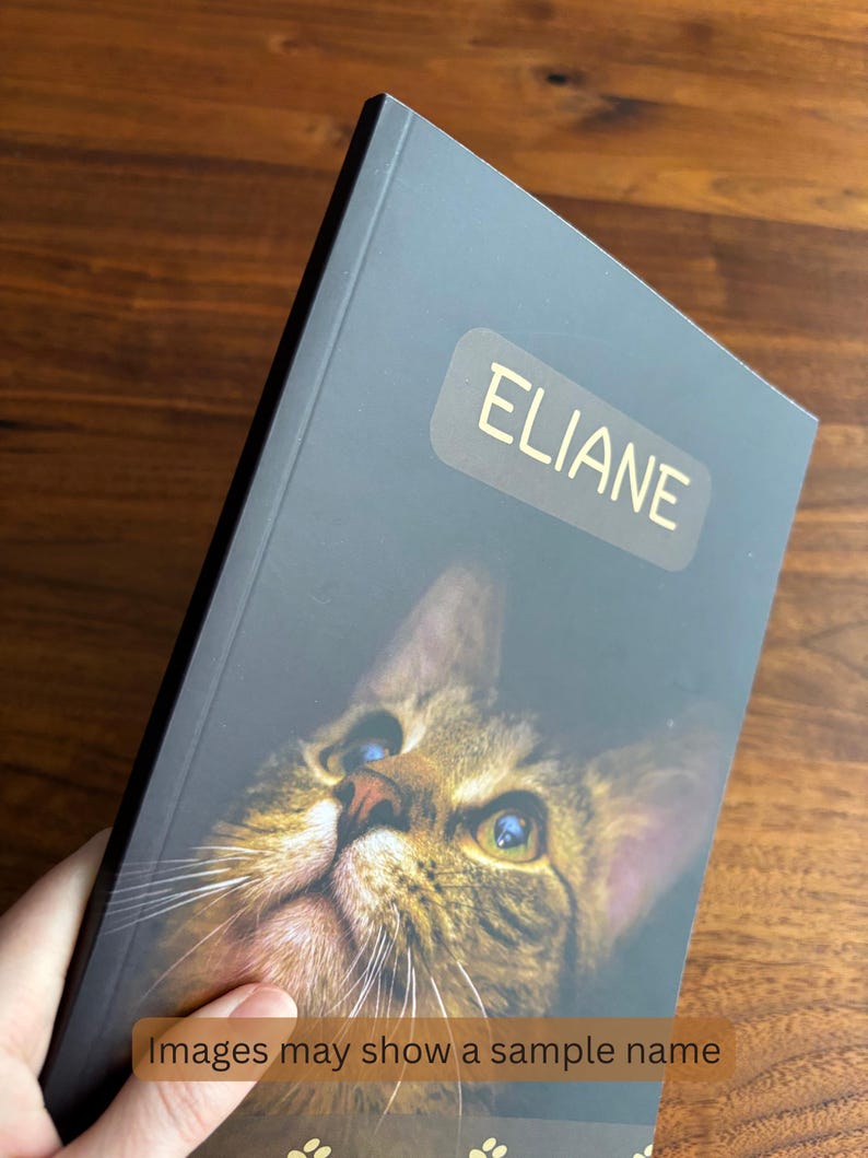 May include: A personalized notebook featuring a close-up image of a cat's face with blue and green eyes. The name "ELIANE" is printed in gold on the cover. The text "Images may show a sample name" is also visible.