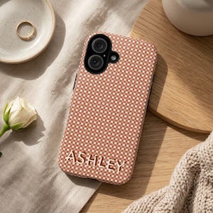 May include: A rectangular mobile phone case featuring a small-scale checkered pattern in shades of terracotta and white. The name ASHLEY is embossed in white capital letters across the lower portion of the case, which has a dual-lens camera cutout.