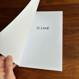 May include: A white paper with the name "ELIANE" printed in black. The page is being turned, revealing a blank page. The image is on a dark wood surface. The bottom of the image has the text "Images may show a sample name".