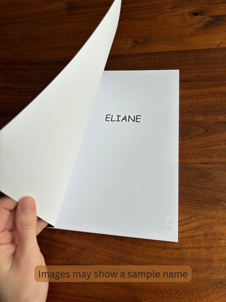 May include: A white paper with the name "ELIANE" printed in black. The page is being turned, revealing a blank page. The image is on a dark wood surface. The bottom of the image has the text "Images may show a sample name".