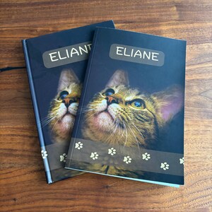 May include: Two personalized notebooks with a cat portrait on the cover. The notebooks have the name "ELIANE" printed on a label. The bottom of the cover features a paw print design. The image text says "Images may show a sample name."