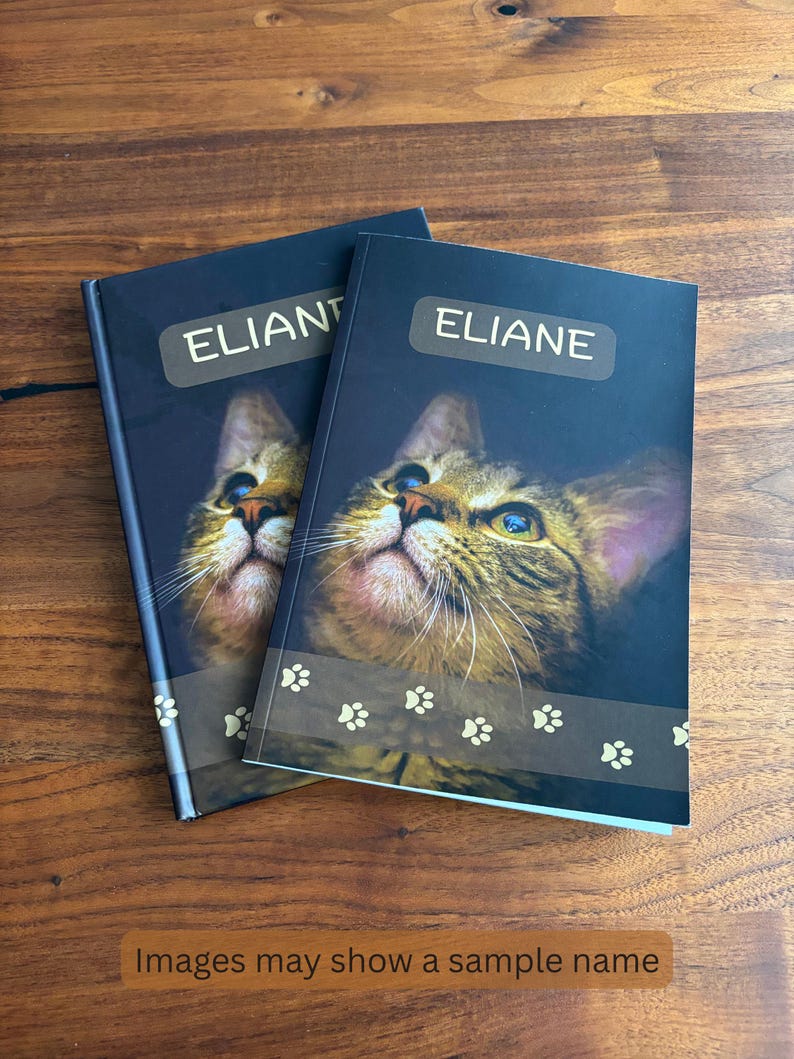 May include: Two personalized notebooks with a cat portrait on the cover. The notebooks have the name "ELIANE" printed on a label. The bottom of the cover features a paw print design. The image text says "Images may show a sample name."