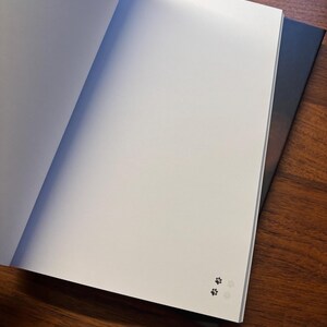May include: An open notebook with blank white pages, featuring a small paw print design in the corner. The notebook is resting on top of another closed notebook with a dark cover, all on a wooden surface.