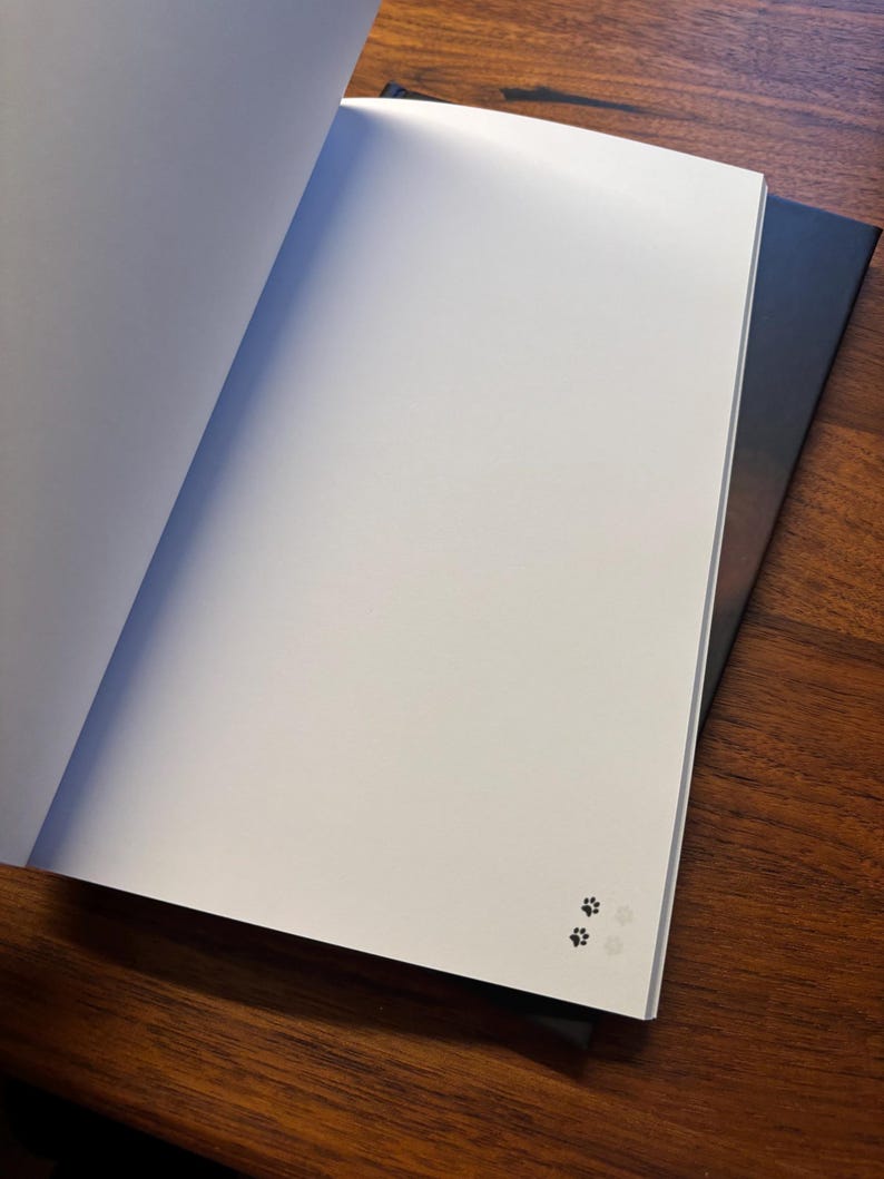 May include: An open notebook with blank white pages, featuring a small paw print design in the corner. The notebook is resting on top of another closed notebook with a dark cover, all on a wooden surface.