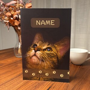 May include: A black notebook featuring a close-up of a tabby cat's face, with the word "NAME" in a gold rectangle at the top. A brown paw print border is at the bottom. A white mug and dried flowers are in the background.