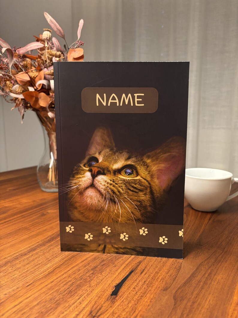 May include: A black notebook featuring a close-up of a tabby cat's face, with the word "NAME" in a gold rectangle at the top. A brown paw print border is at the bottom. A white mug and dried flowers are in the background.