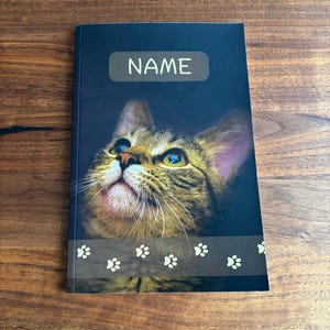 May include: A notebook with a dark blue cover featuring a close-up of a tabby cat's face. The word "NAME" is printed in white on a light brown rectangle. A paw print design is at the bottom.