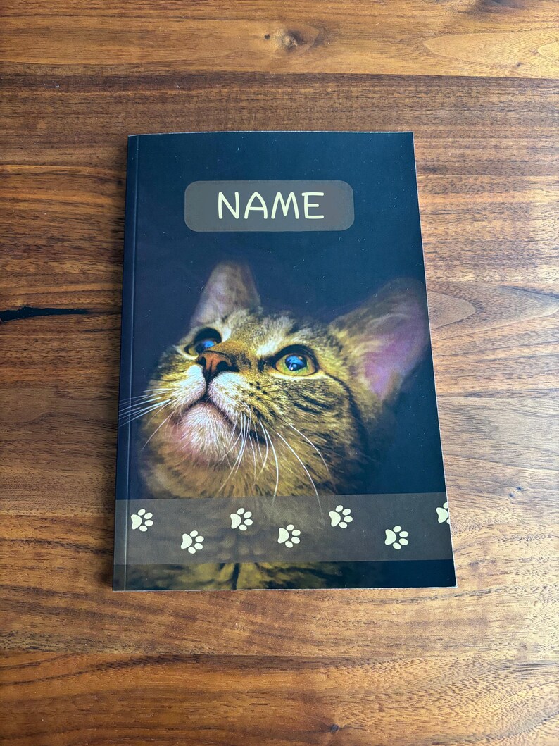 May include: A notebook with a dark blue cover featuring a close-up of a tabby cat's face. The word "NAME" is printed in white on a light brown rectangle. A paw print design is at the bottom.