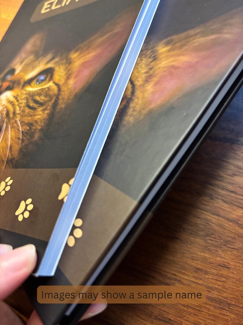 May include: A close-up of a book with a cat's face on the cover. The cover is dark with gold accents and paw prints. A light blue plastic strip is visible on the side. The text "Images may show a sample name" is at the bottom.