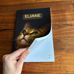 May include: A notebook with the name "ELIANE" in gold lettering on the cover. The cover features a close-up image of a cat's face. The notebook is open, revealing white pages. The text "Images may show a sample name" is at the bottom.