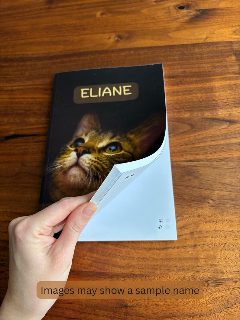 May include: A notebook with the name "ELIANE" in gold lettering on the cover. The cover features a close-up image of a cat's face. The notebook is open, revealing white pages. The text "Images may show a sample name" is at the bottom.