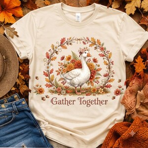 Peut inclure: Un t-shirt à manches courtes couleur crème avec un motif central représentant une oie blanche portant une couronne de fleurs, entourée d'une couronne automnale de feuilles, de glands et de fleurs, avec le texte Gather Together imprimé.