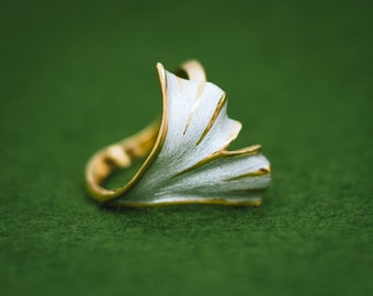 May include: A gold ring with a white enamel ginkgo leaf design. The ring is on a green surface.