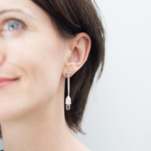 May include: Silver-toned drop earrings. Each earring features a small star-shaped stud, a long, slender bar, and a clear, teardrop-shaped crystal pendant. The earrings are worn by a person with brown hair.