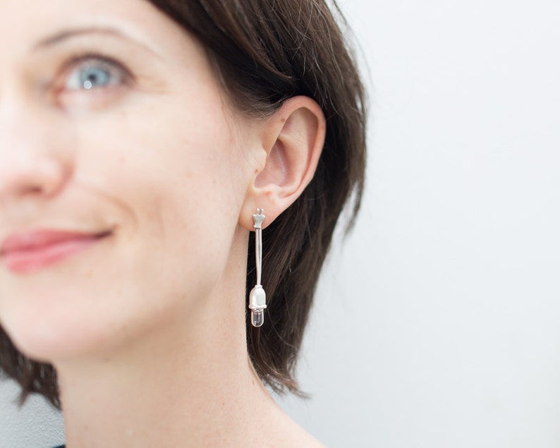 May include: Silver-toned drop earrings. Each earring features a small star-shaped stud, a long, slender bar, and a clear, teardrop-shaped crystal pendant. The earrings are worn by a person with brown hair.