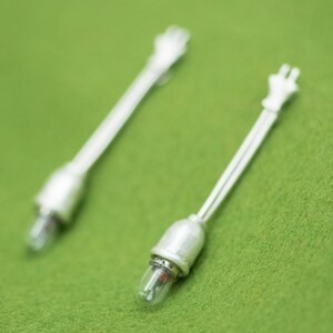 May include: Two miniature silver-colored light bulbs with clear glass bulbs and white wiring, resting on a green surface. The bulbs have a plug-like design at the end of the wire.
