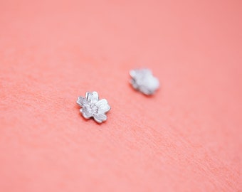 May include: Two silver flower-shaped earrings on a pink surface.