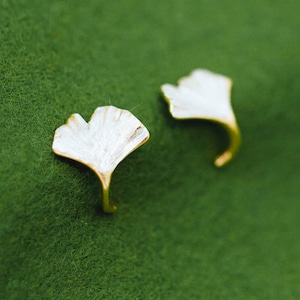 Könnte beinhalten: Ein Paar goldfarbene Ohrringe mit weißen Ginkgoblatt-Designs. Die Blätter sind detailliert und haben eine leicht strukturierte Oberfläche.