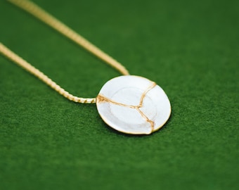 Pode incluir: Um colar de corrente de ouro com um pingente de cerâmica branca. O pingente tem um design dourado que parece rachaduras na cerâmica.