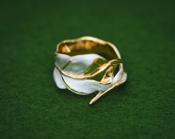 May include: A silver and gold ring with a leaf design. The ring is on a green background.