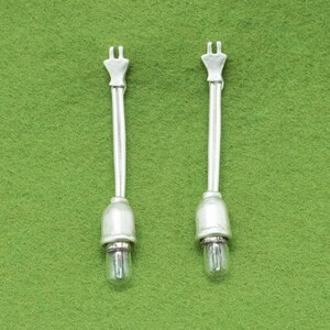 May include: Two silver-toned light bulb fixtures with a clear glass bulb at the base. Each fixture has a long, thin stem and a decorative top. The background is a solid green color.