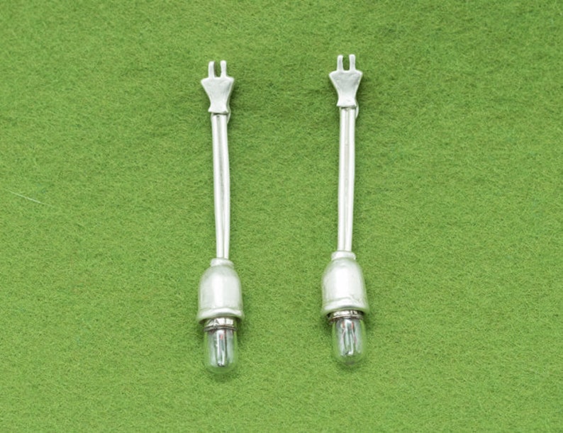 May include: Two silver-toned light bulb fixtures with a clear glass bulb at the base. Each fixture has a long, thin stem and a decorative top. The background is a solid green color.