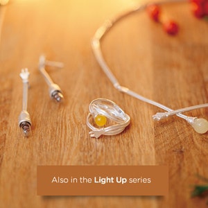 May include: A collection of silver-toned jewelry pieces, including a necklace, earrings, a ring with a yellow stone, and decorative sticks. The items are arranged on a wooden surface with a warm, inviting glow. The text "Also in the Light Up series" is visible.