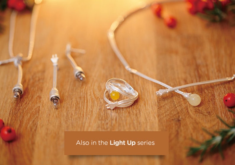 May include: A collection of silver-toned jewelry pieces, including a necklace, earrings, a ring with a yellow stone, and decorative sticks. The items are arranged on a wooden surface with a warm, inviting glow. The text "Also in the Light Up series" is visible.