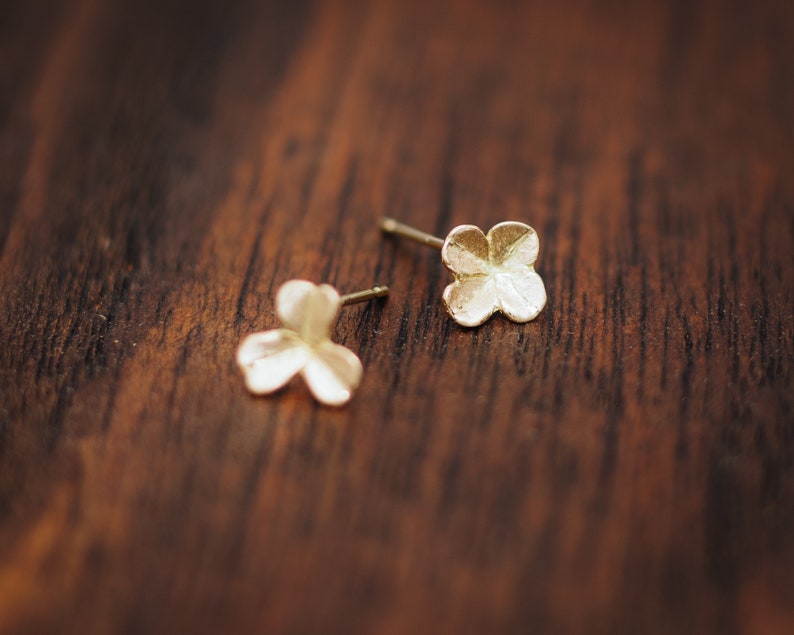 Op de afbeelding: Twee kleine, goudkleurige oorstekers. Elke oorbel heeft een vierbladige bloemvorm. De oorbellen zijn geplaatst op een donker houten oppervlak. De oorknopjes zijn delicaat en lijken van metaal te zijn.
