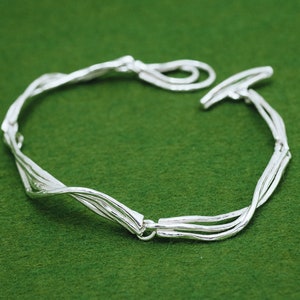Op de afbeelding: Een zilveren armband met een gedraaid, geweven ontwerp. De armband is gemaakt van meerdere zilveren draden die met elkaar zijn verweven om een uniek en ingewikkeld patroon te cre&euml;ren.