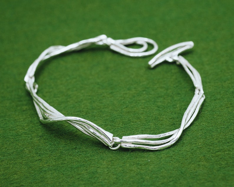 Op de afbeelding: Een zilveren armband met een gedraaid, geweven ontwerp. De armband is gemaakt van meerdere zilveren draden die met elkaar zijn verweven om een uniek en ingewikkeld patroon te cre&euml;ren.