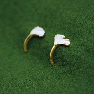 May include: A pair of gold earrings with white ginkgo leaf designs. The earrings are on a green background.