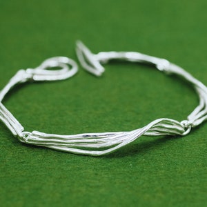 Op de afbeelding: Een zilveren armband met een geweven ontwerp. De armband is gemaakt van meerdere zilveren draden die met elkaar zijn gedraaid om een uniek en ingewikkeld patroon te cre&euml;ren.