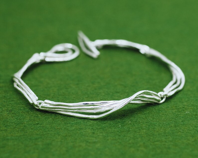 Op de afbeelding: Een zilveren armband met een geweven ontwerp. De armband is gemaakt van meerdere zilveren draden die met elkaar zijn gedraaid om een uniek en ingewikkeld patroon te cre&euml;ren.