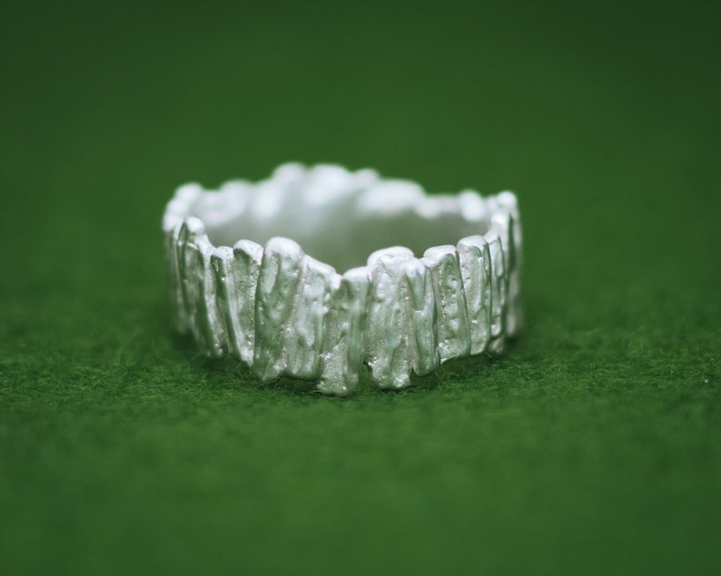 May include: A silver ring with a textured, organic design. The ring is made of a series of vertical, uneven shapes that resemble a rough, natural surface.