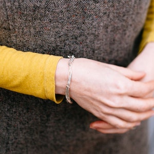 Op de afbeelding: Een zilveren armband met een geweven ontwerp. De armband wordt om de pols gedragen.