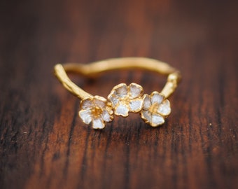 May include: A gold ring with three white flower accents. The ring is made of a textured metal and has a delicate design.