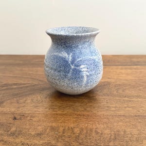 May include: A small, round, blue and white speckled ceramic vase with a wide opening. The vase has a slightly flared rim and a textured surface. The blue colour is a soft, muted shade, and the white speckles add visual interest.