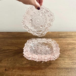 May include: Two pink glass dishes with detailed starburst and floral patterns. One dish is held above the other, which is on a wooden surface. The dishes have scalloped edges and are likely decorative or for serving.