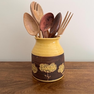 May include: A yellow and brown ceramic jar containing wooden spoons and forks. The jar is decorated with the word "SUGAR". The jar is placed on a wooden surface.