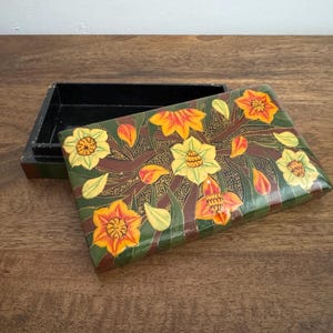 May include: A rectangular wooden box with a hinged lid, painted with a floral design. The lid features yellow and orange flowers, green leaves, and brown stems on a dark green background. The box is open, revealing a black interior.