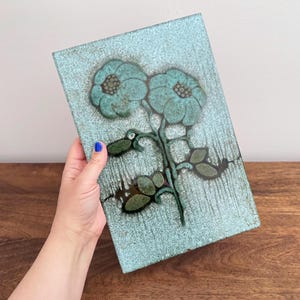May include: A rectangular ceramic wall plaque featuring a textured light blue and turquoise glaze. The surface displays a dark brown and green floral motif with two prominent blossoms and several leaves, accented by vertical drip-like patterns.