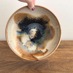 May include: A handmade ceramic bowl with a unique glaze. The bowl has a cream-coloured exterior and a centre with brown, blue, and gold hues. The bowl is held by a hand.