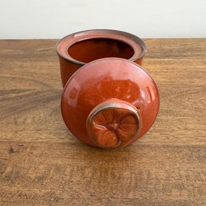 May include: An open, orange ceramic sugar bowl with a matching lid. The lid is round with a decorative floral design on the handle. The bowl and lid have a glossy finish and sit on a wooden surface.